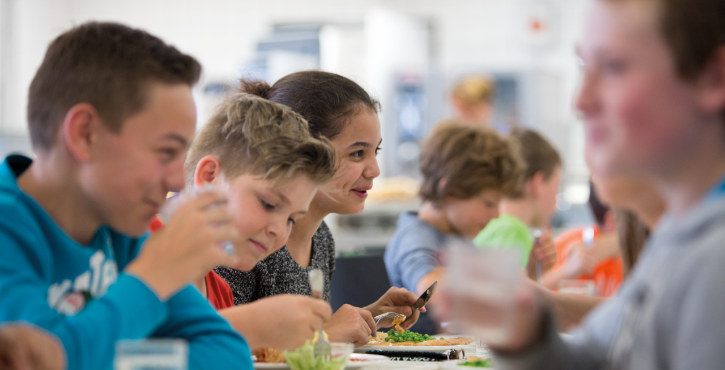 A group of children enjoying lunch in a school cafeteria, smiling and engaged while eating vegetables and other dishes.