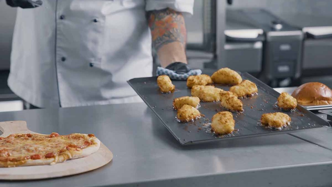 A chef takes a tray of crispy food from the oven, with a pizza on a wooden board in the foreground.