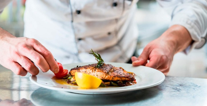 A chef garnishing a plate of grilled salmon with cherry tomatoes and lemon on a restaurant table.