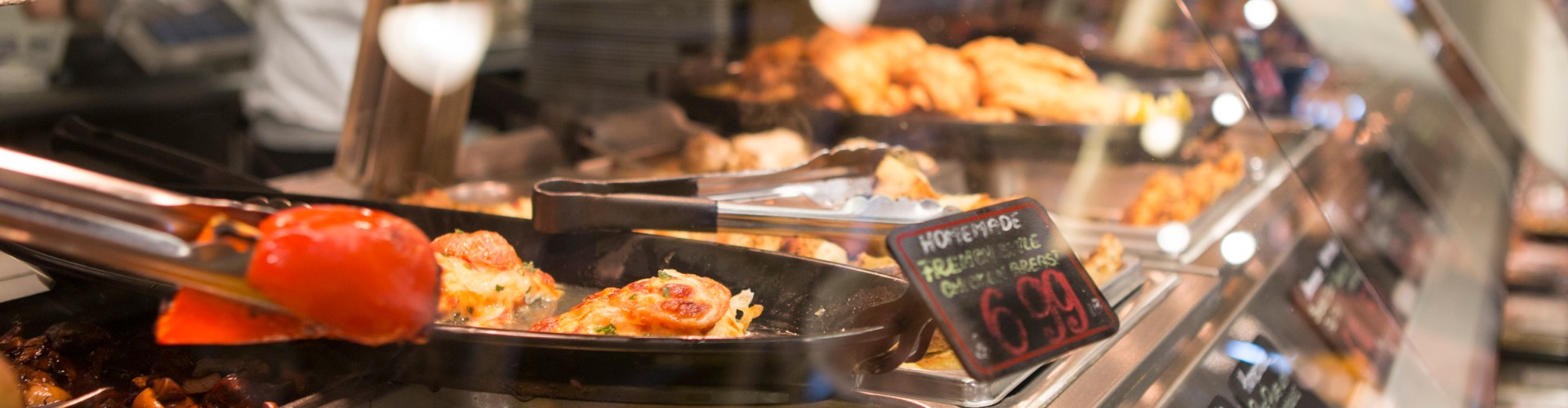 A close-up of a supermarket food display with various prepared meals and prices, showcasing vibrant food items and customer interaction.