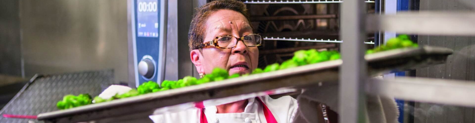 A chef wearing glasses and an apron is removing trays of broccoli from a Rational oven in a kitchen setting.