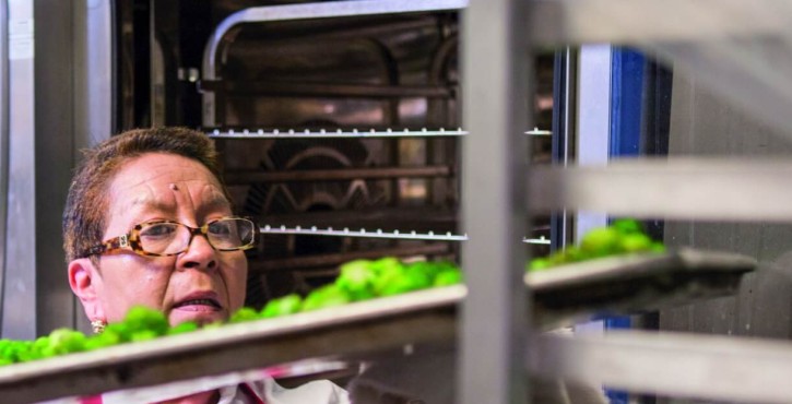 Una chef con gafas y un delantal está sacando bandejas de brócoli de un horno Rational en un entorno de cocina.