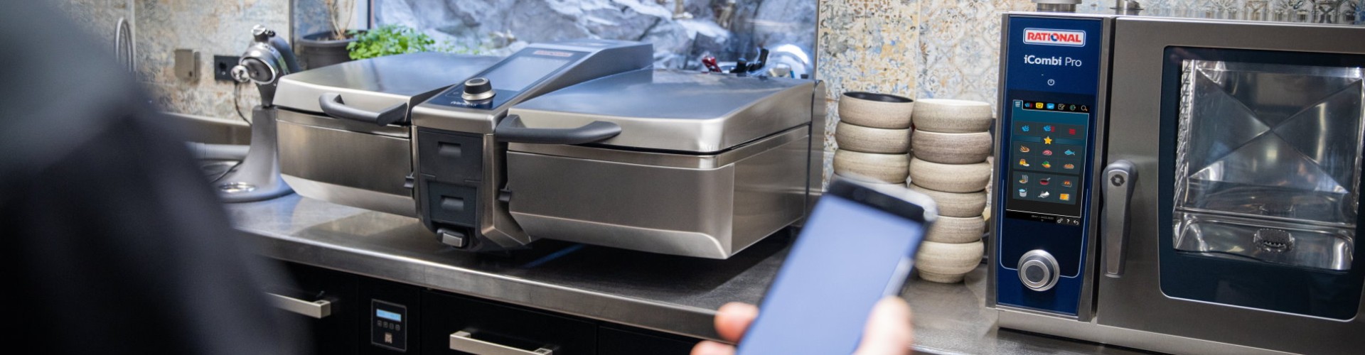 Person controlling a modern kitchen with a Rational iVario and iCombi Pro using a smartphone.