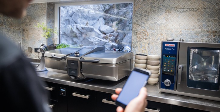 Person controlling a modern kitchen with a Rational iVario and iCombi Pro using a smartphone.