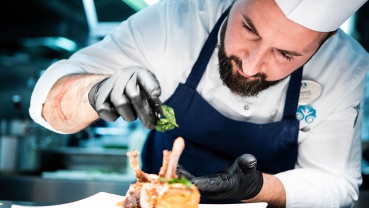 A chef artfully garnishes a plate with a portion of meat and fresh herbs in a restaurant kitchen.