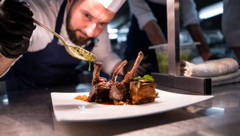 A chef garnishing a plate of lamb chops with sauce, showcasing the dish's intricate presentation in a professional kitchen.