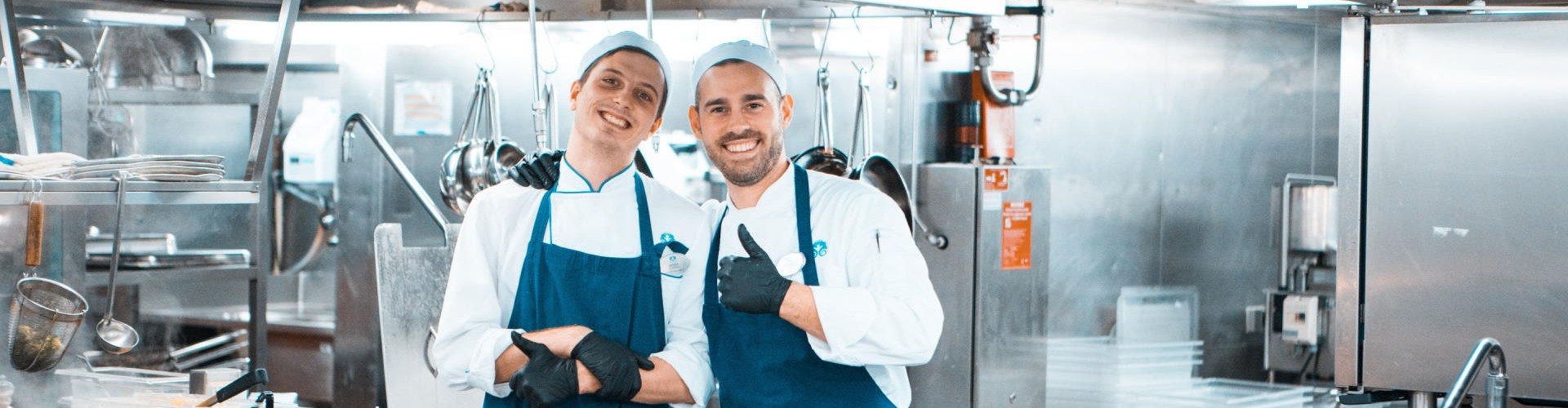 Two smiling chefs posing in a Ship‘s galley, wearing blue aprons and gloves, with kitchen equipment in the background.