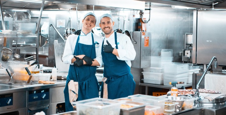 Two smiling chefs posing in a Ship's galley, wearing blue aprons and gloves, with kitchen equipment in the background.