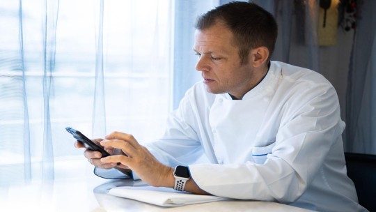 Chef in a white uniform sitting by a window, using a smartphone and looking focused.