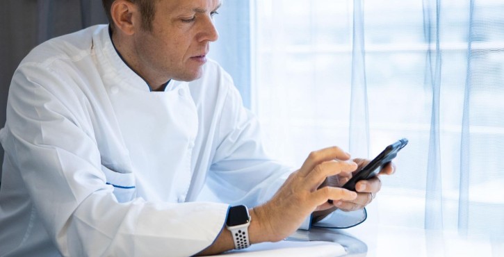 Un chef en uniforme blanc examine son smartphone tout en étant assis à une table.