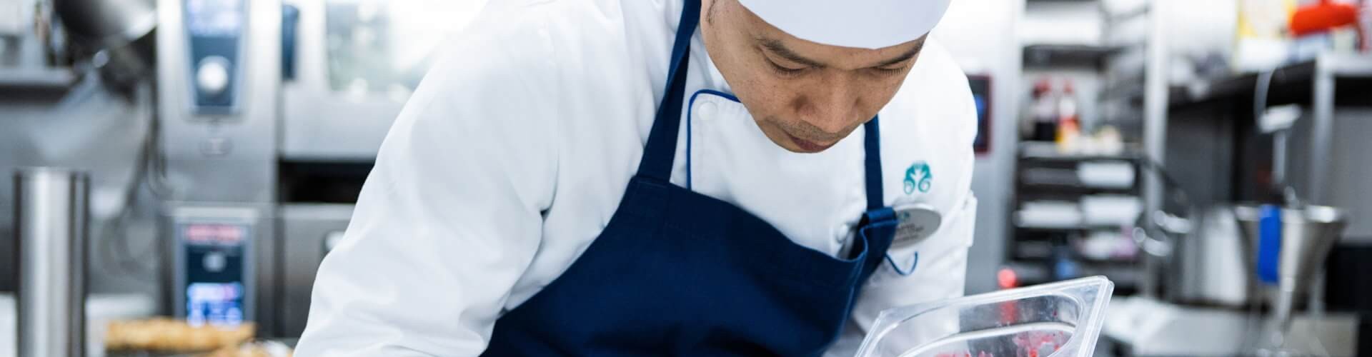 A chef in a kitchen carefully plating a dessert, adding berry sauce next to a slice of cake on white plates.