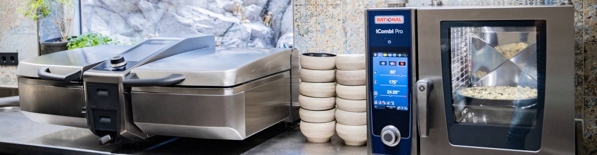 Una cocina con un horno Rational iCombi Pro y un electrodoméstico Vario Pro en una encimera de acero inoxidable, con tazones apilados al fondo.