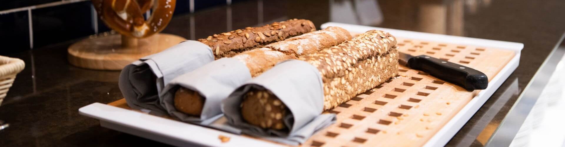 Assortment of seeded baguettes and soft pretzels on a breakfast buffet counter.