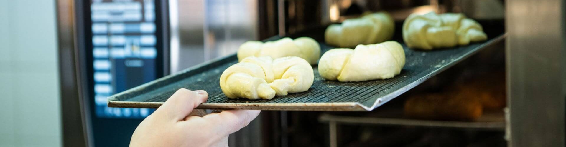 A person placing uncooked croissants on a tray into a Rational oven with a digital display in a commercial kitchen.