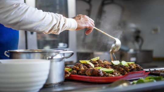 Chef pouring sauce over roasted meat with limes in a professional kitchen.