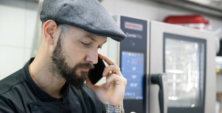 A chef wearing a gray cap talks on the phone while working in a kitchen, with an iCombi Pro cooking system visible in the background.