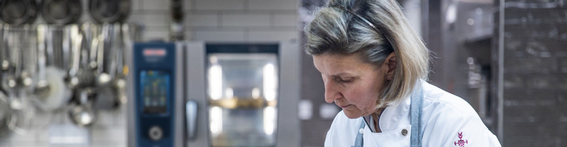 Un chef en una cocina profesional está preparando un platillo, colocando ingredientes en una bandeja. El chef lleva una chaqueta blanca y un delantal gris, con un horno iCombi Pro visible al fondo.