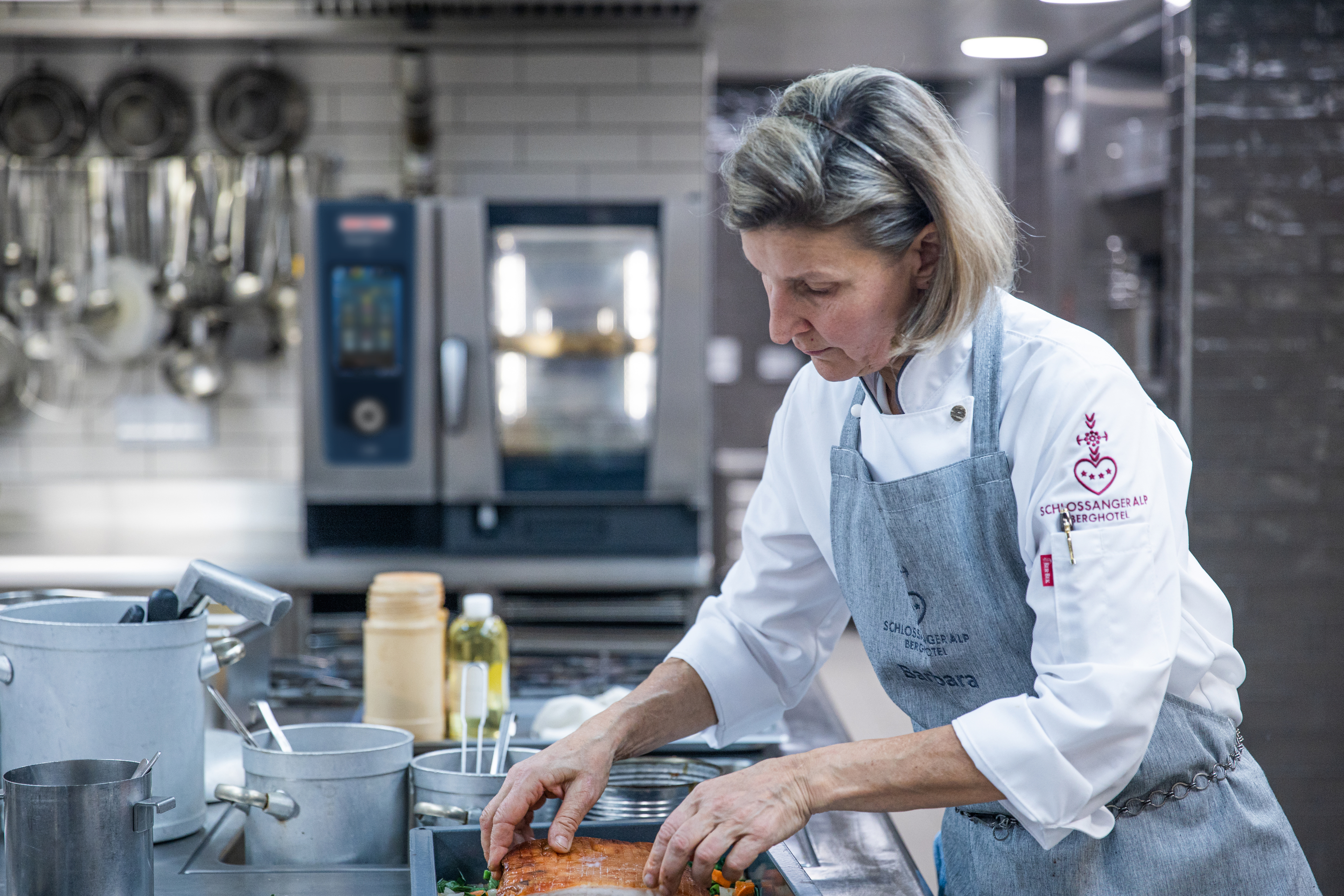 A chef in a professional kitchen is preparing a dish, placing ingredients into a tray. The chef is wearing a white coat and a gray apron, with an iCombi Pro oven visible in the background.