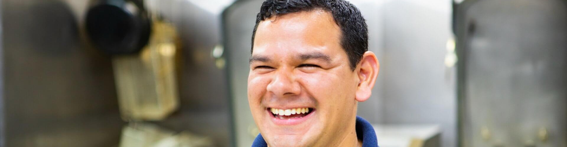A smiling male chef wearing a navy shirt and white apron stands in a kitchen filled with cooking equipment.