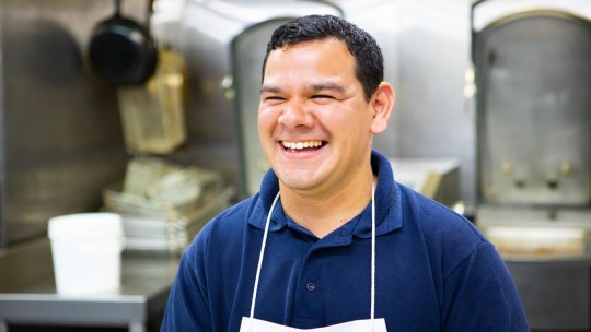 A smiling male chef wearing a navy shirt and white apron stands in a kitchen filled with cooking equipment.