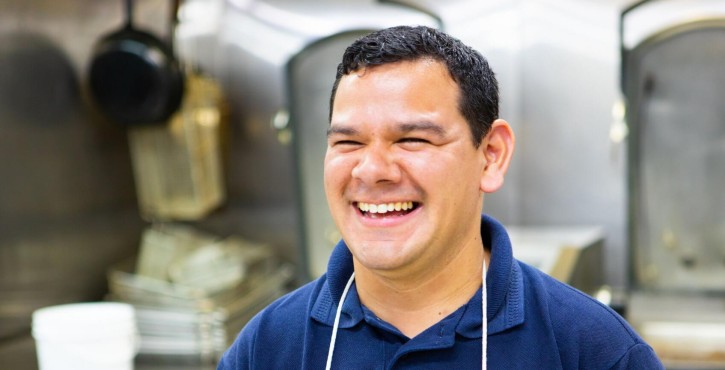 A smiling male chef wearing a navy shirt and white apron stands in a kitchen filled with cooking equipment.