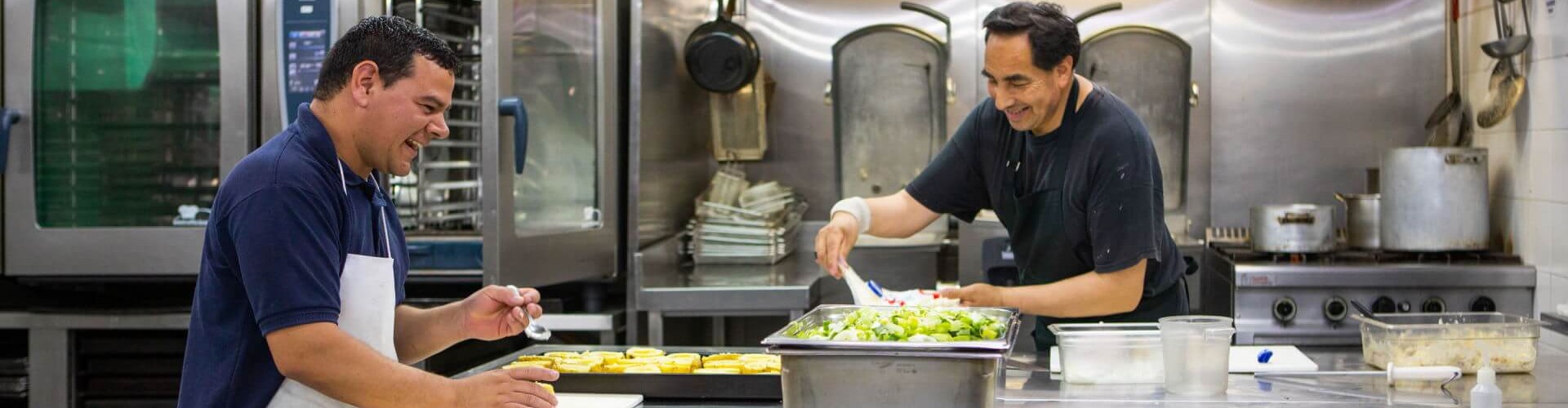 Two chefs working together in a kitchen, one preparing a dish while the other is arranging food on a plate, with Rational ovens in the background.