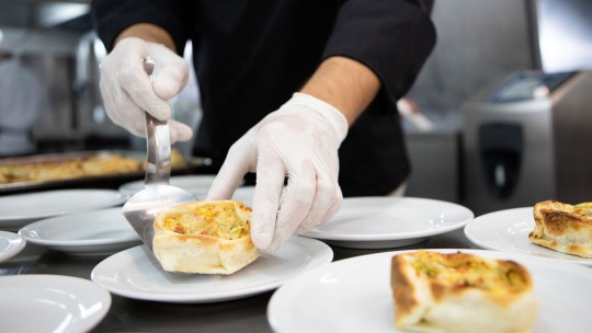 Chef plating a vegetable-filled pastry on a white plate in a busy kitchen.