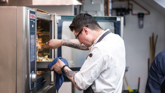 A chef pulls a tray of food from a RATIONAL oven while another person stands in the background.