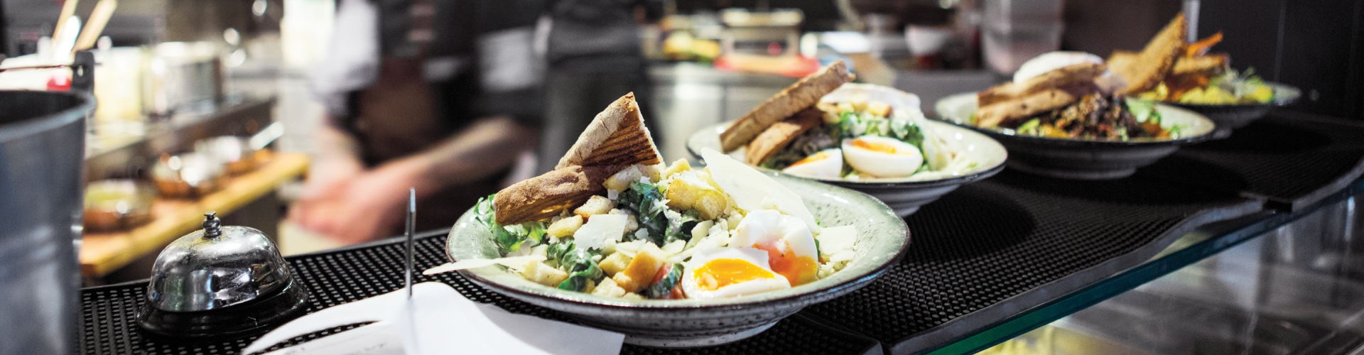 Several plates of food, including salad and eggs, are ready to be served in a busy restaurant kitchen, with chefs in the background.