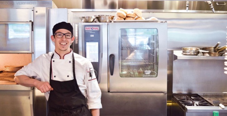 A smiling chef standing next to a Rational cooking appliance in a professional kitchen, with various kitchen equipment in the background.