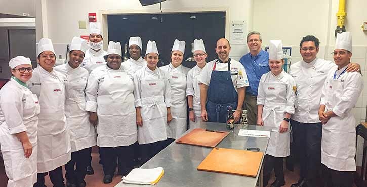 A group photo of culinary students and instructors posing together in a kitchen setting, all wearing chef uniforms and hats.