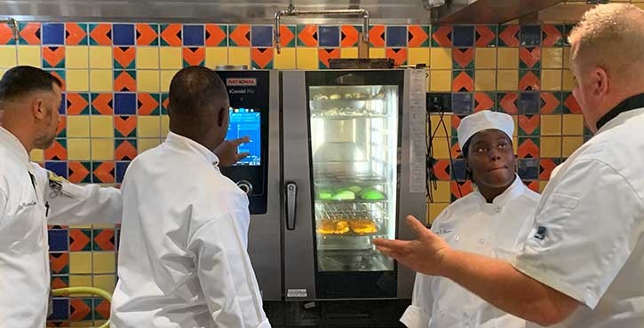 A group of culinary students observing an instructor as they interact with a Rational iCombi Pro oven's touchscreen in a colorful kitchen.