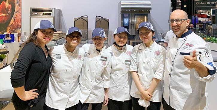 A group of culinary students and an instructor posing together in a kitchen setting, all wearing chef coats and caps, smiling at the camera.