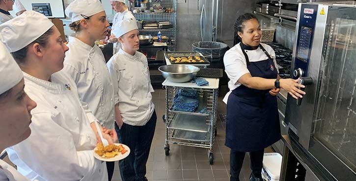 A culinary instructor demonstrates how to operate a Rational iCombi oven while students watch attentively in a professional kitchen.