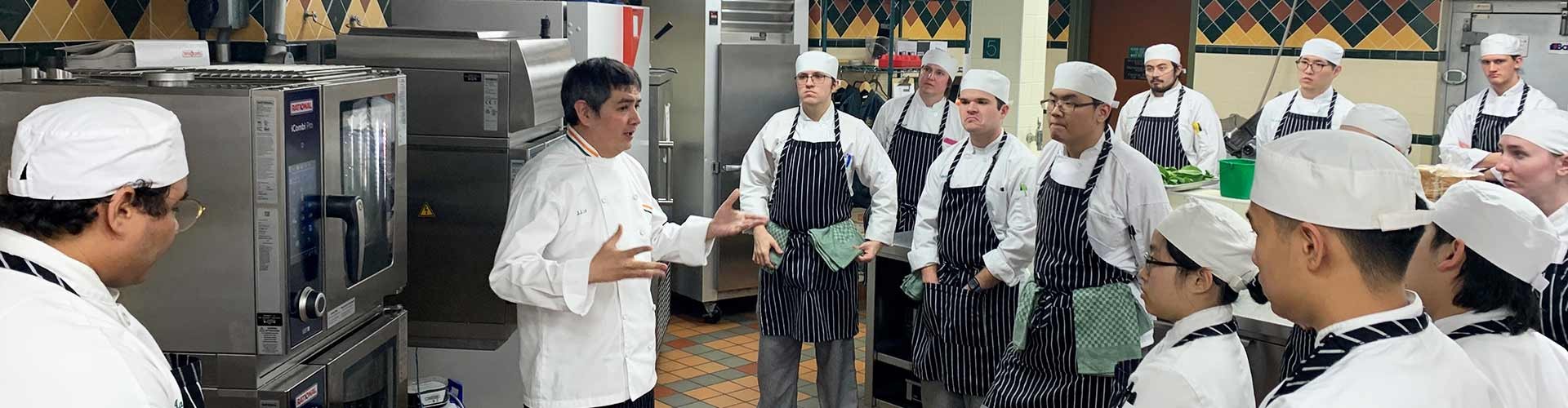A group of culinary students attentively listening to an instructor in a kitchen, with a Rational iCombi Pro oven in the background.