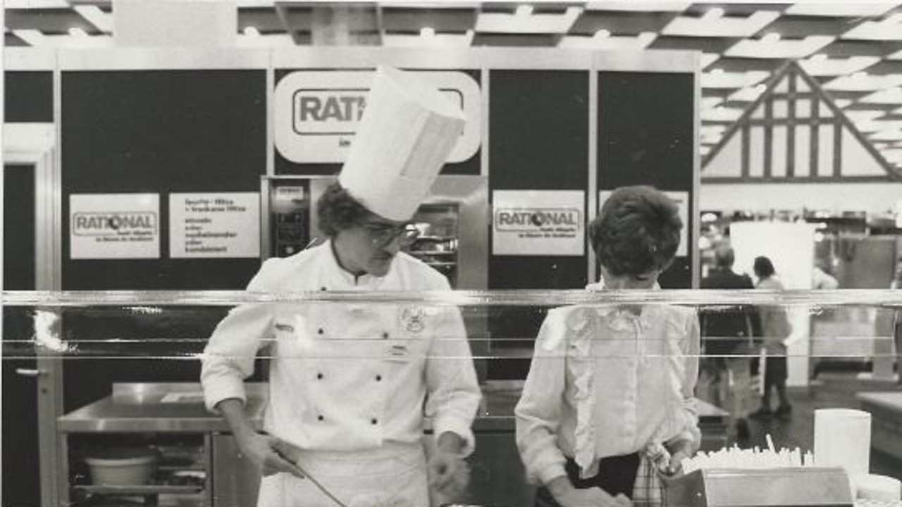 A black-and-white photo of two individuals at a Rational booth during a trade show, one in a chef‘s uniform and hat, preparing food.