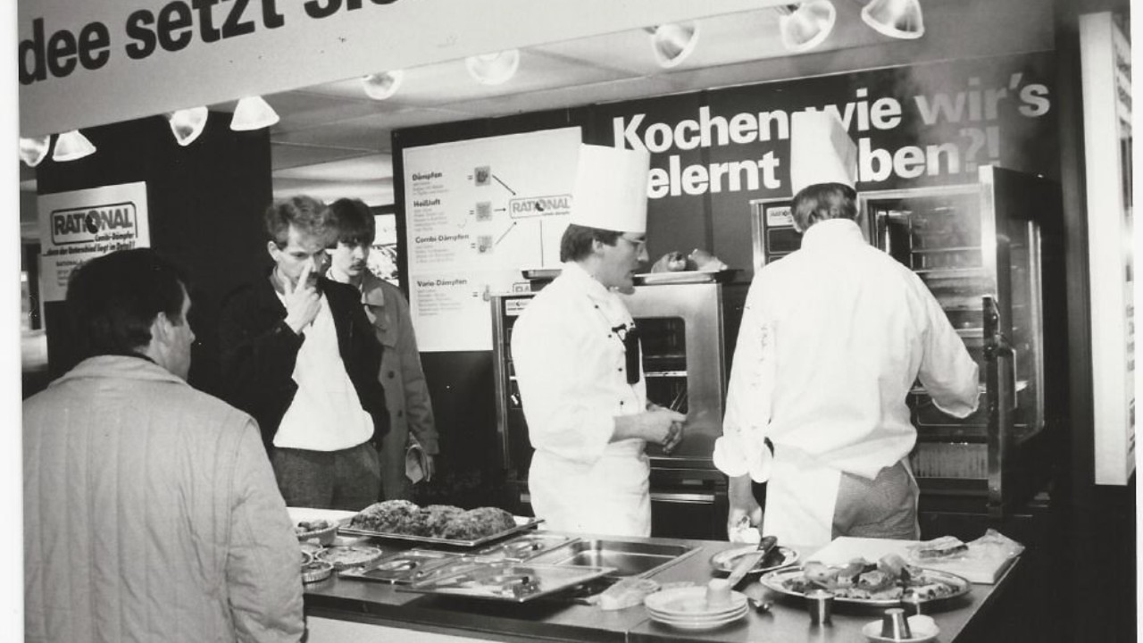 A black-and-white photo of a Rational trade show booth with chefs demonstrating cooking to an audience, featuring various dishes on display.