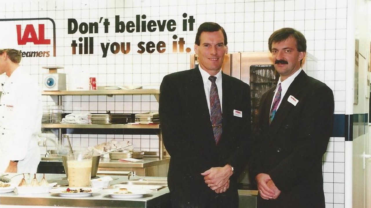 A trade show photo from 1994 in the USA featuring two men in suits standing in front of a kitchen setup, with a chef preparing food in the background.