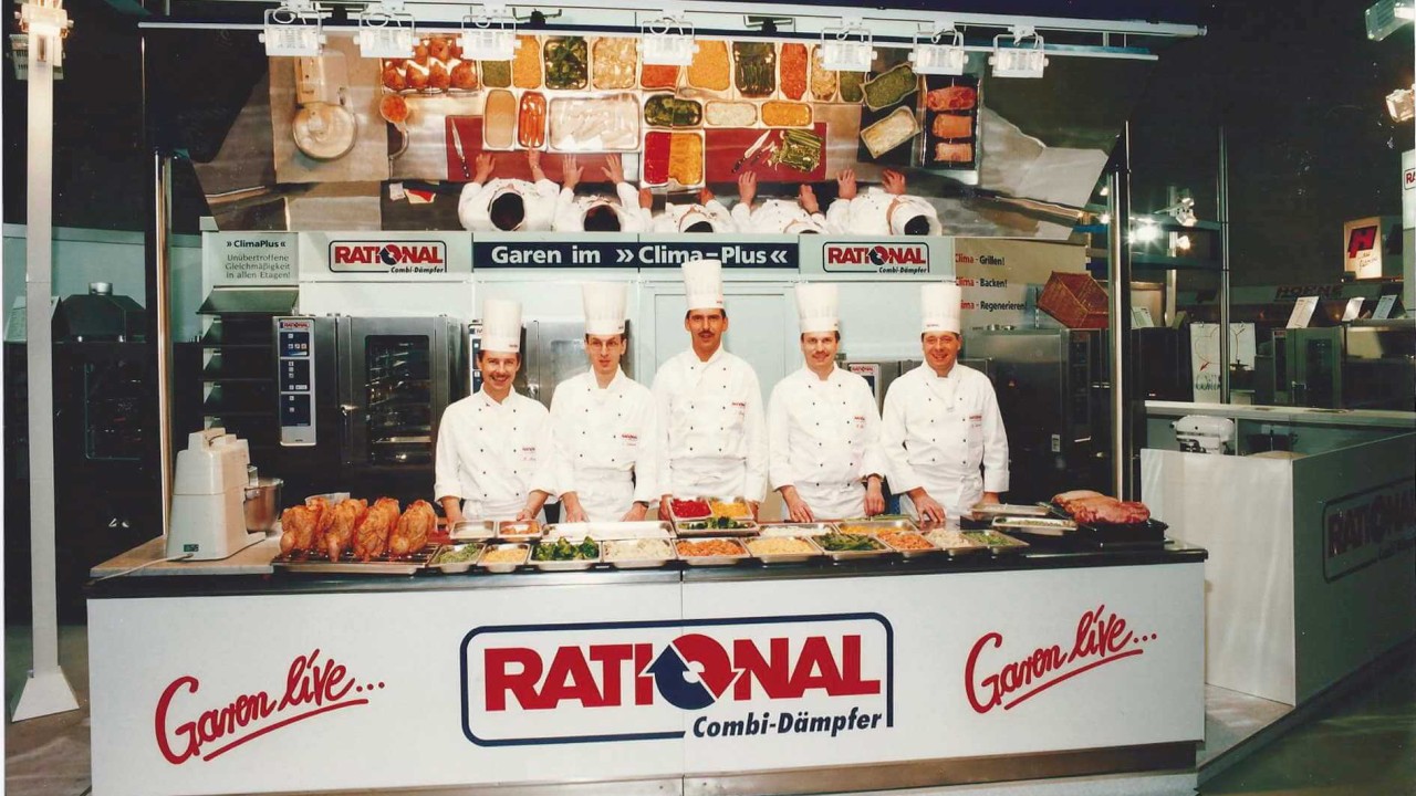 A 1995 trade show photo featuring five chefs in white uniforms standing behind a display of food, with cooking equipment and the Rational logo in the background.