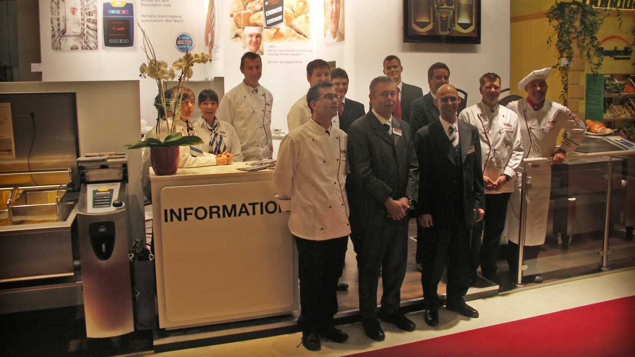 A 2009 trade show photo featuring a group of chefs and representatives standing in front of a display showcasing cooking equipment, with promotional signage in German.