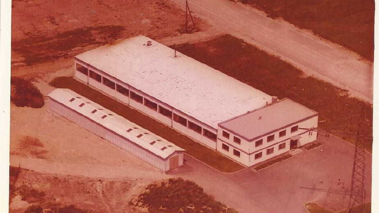 An aerial view of a factory building from 1973, showing a large main building with a flat roof and an adjacent smaller structure, surrounded by dirt paths and greenery.
