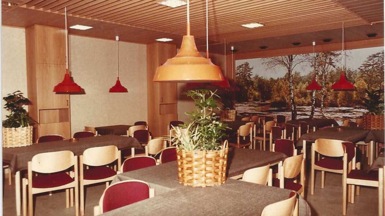 An interior view of a cafeteria from 1984, featuring wooden tables with gray tablecloths, red pendant lamps, and a background mural of a forest landscape.