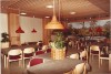 An interior view of a cafeteria from 1984, featuring wooden tables with gray tablecloths, red pendant lamps, and a background mural of a forest landscape.