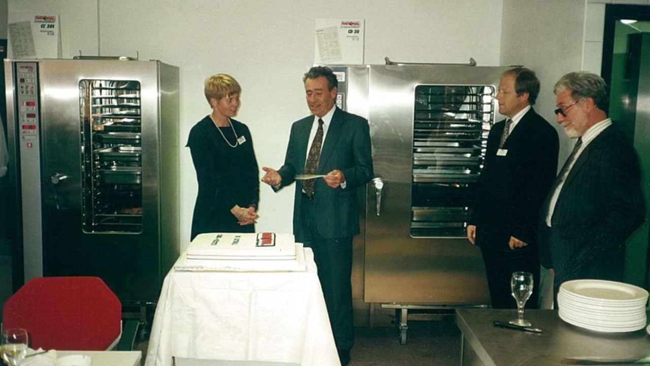 A celebration at the opening of a subsidiary company in 1991, with a cake and attendees in formal attire near Rational ovens.