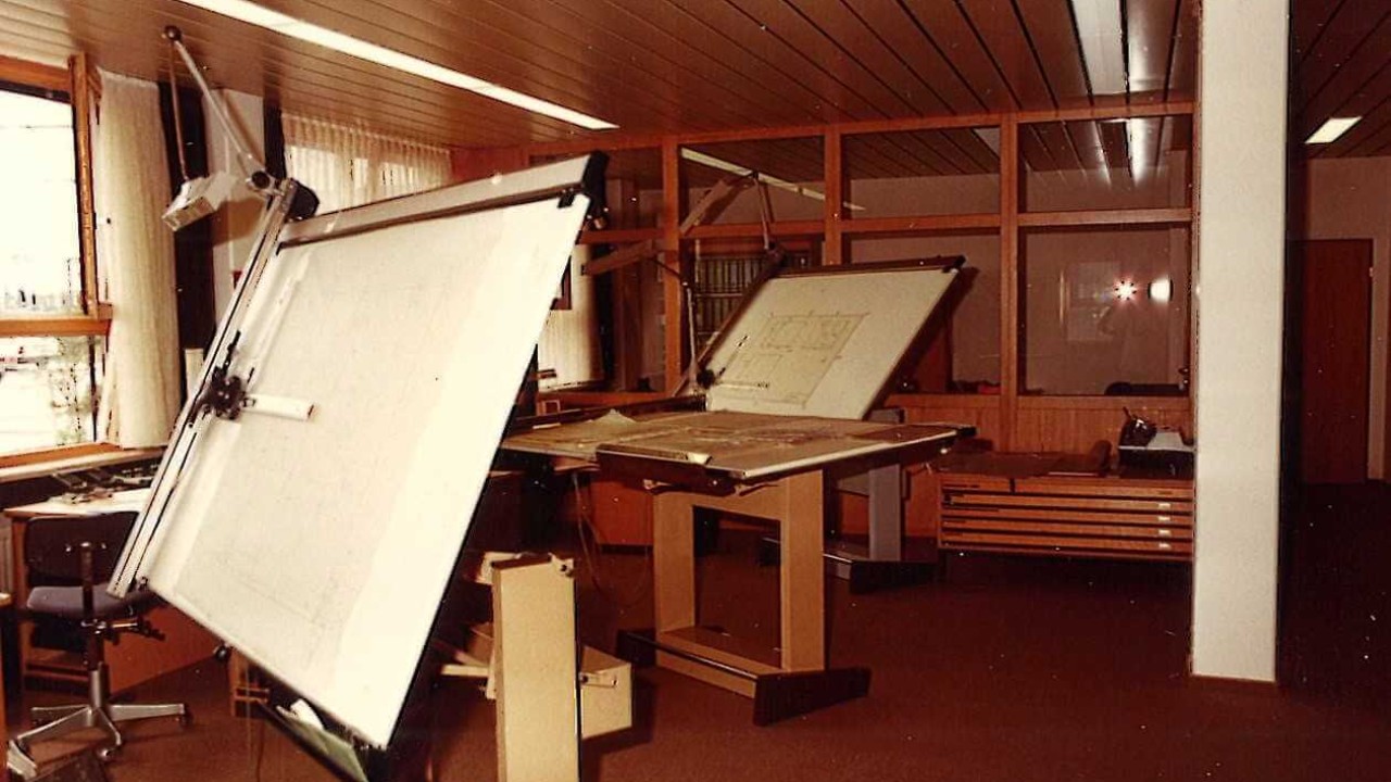 An interior view of an engineering drawing room from the past, featuring drafting tables, a rolling chair, and bright overhead lighting.