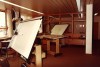 An interior view of an engineering drawing room from the past, featuring drafting tables, a rolling chair, and bright overhead lighting.
