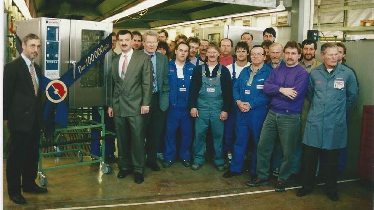 Group photo of employees celebrating the production of the 100,000th Rational combi-steamer in 1996, with a large ribbon displayed.
