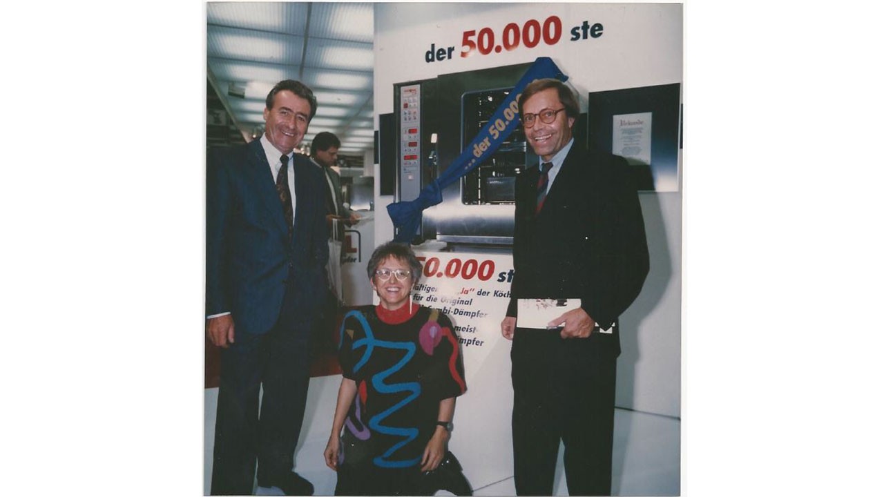 Three people celebrating the sale of the 50,000th Rational combi-steamer in 1991, with a large banner in the background.