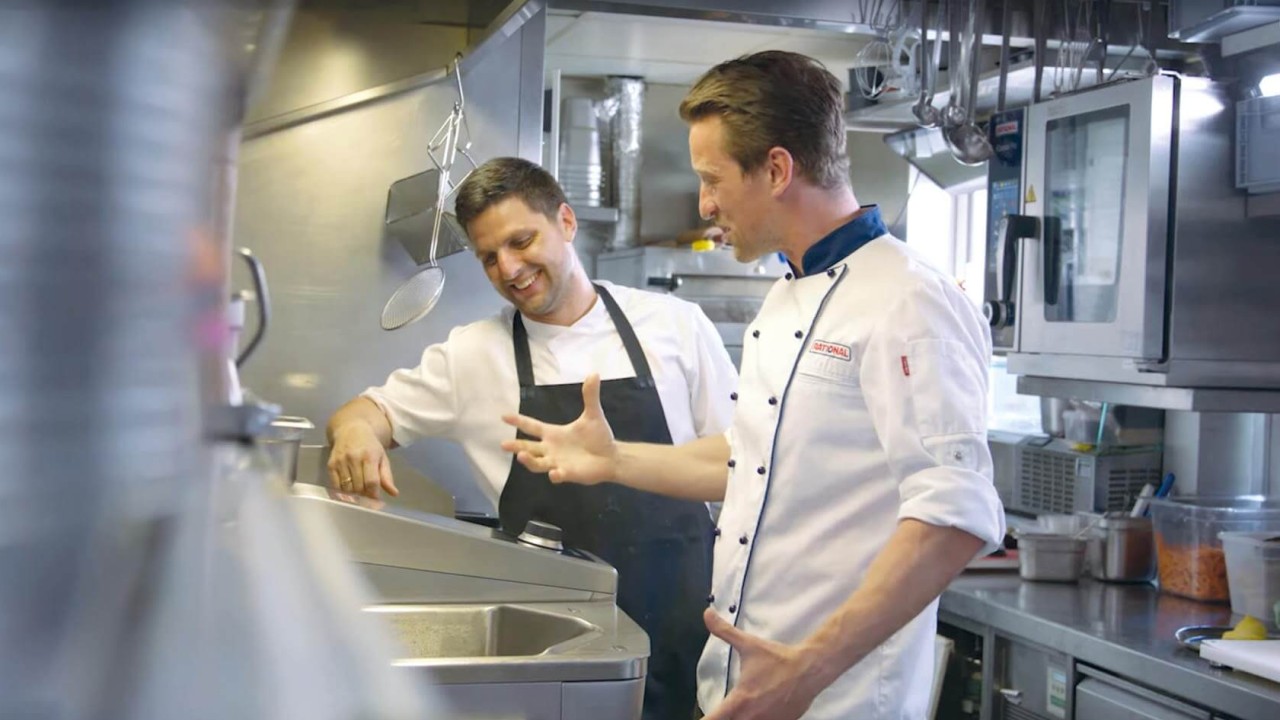 Two chefs in a commercial kitchen, one in a white chef‘s coat and the other in a black apron, discussing food preparation over a iVario Pro.