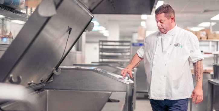 Chef Stefan Pappert in a white uniform operating a large commercial cooking appliance in a professional kitchen setting.